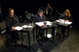 Drei Vorleser sitzen an schwarzen Tischen mit Mikrofonen