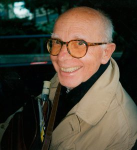 Portraitfoto des Autors auf dem Rücksitz eines Auto mit einer braunen Jacke und einer Tasche umgehongen