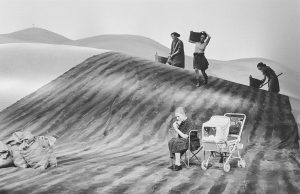 schwarz weiß Foto einer Bühne. Drei Frauen arbeiten im Hintergrund auf einem Feld, eine alte Frau sitzt vornedran mit einem Kinderwagen